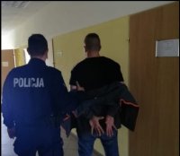 umundurowany policjant stoi z zatrzymanym na korytarzu komendy. są tyłem.zatrzymany ma ręce skute kajdankami