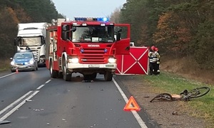 miejsce wypadku drogowego, wozy służb ratowniczych stojące na drodze, obok czerwony parawan, po prawej połamany rower leżący na poboczu
